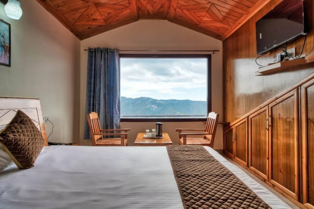 Un dormitorio con una cama grande, dos sillones, una mesita, un televisor montado en la pared y una ventana con vistas a la montaña.