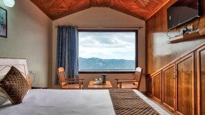 Un dormitorio con una cama grande, dos sillones, una mesita, un televisor montado en la pared y una ventana con vistas a la montaña.