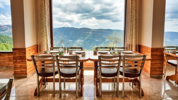 Un comedor con una mesa dispuesta para cuatro, ventanas amplias con cortinas y vistas a montañas.