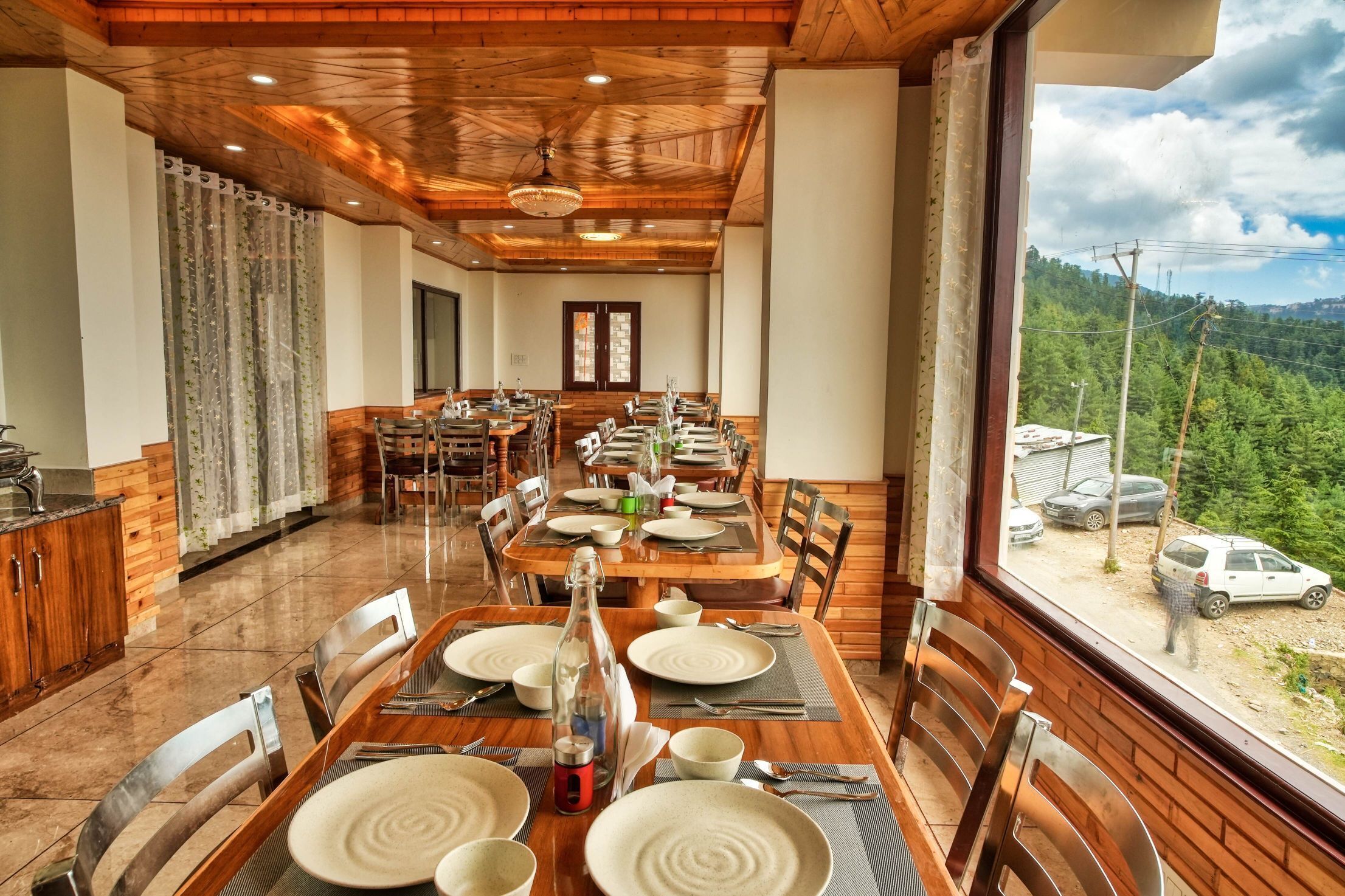 A dining area with wooden tables and chairs, a large window with a view of a forested area, and a door in the background.