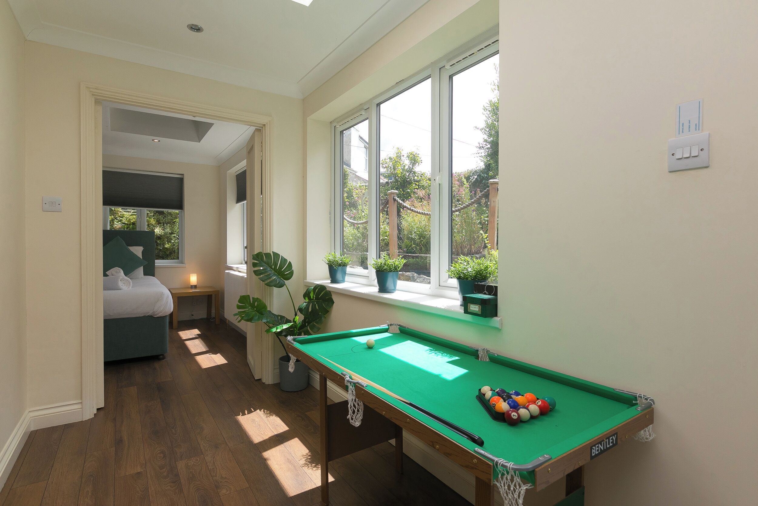 A pool table with a green felt surface, a bed with white bedding, and a window with potted plants.