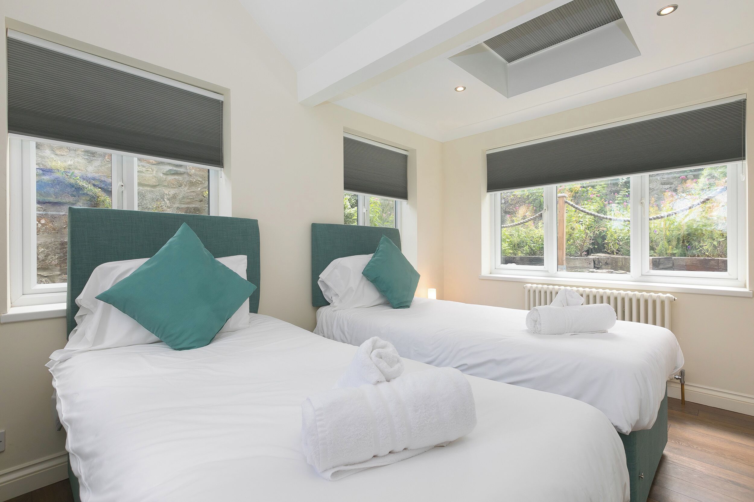 A hotel room with two beds, each with white linens and teal pillows, large windows with blinds, and a skylight.