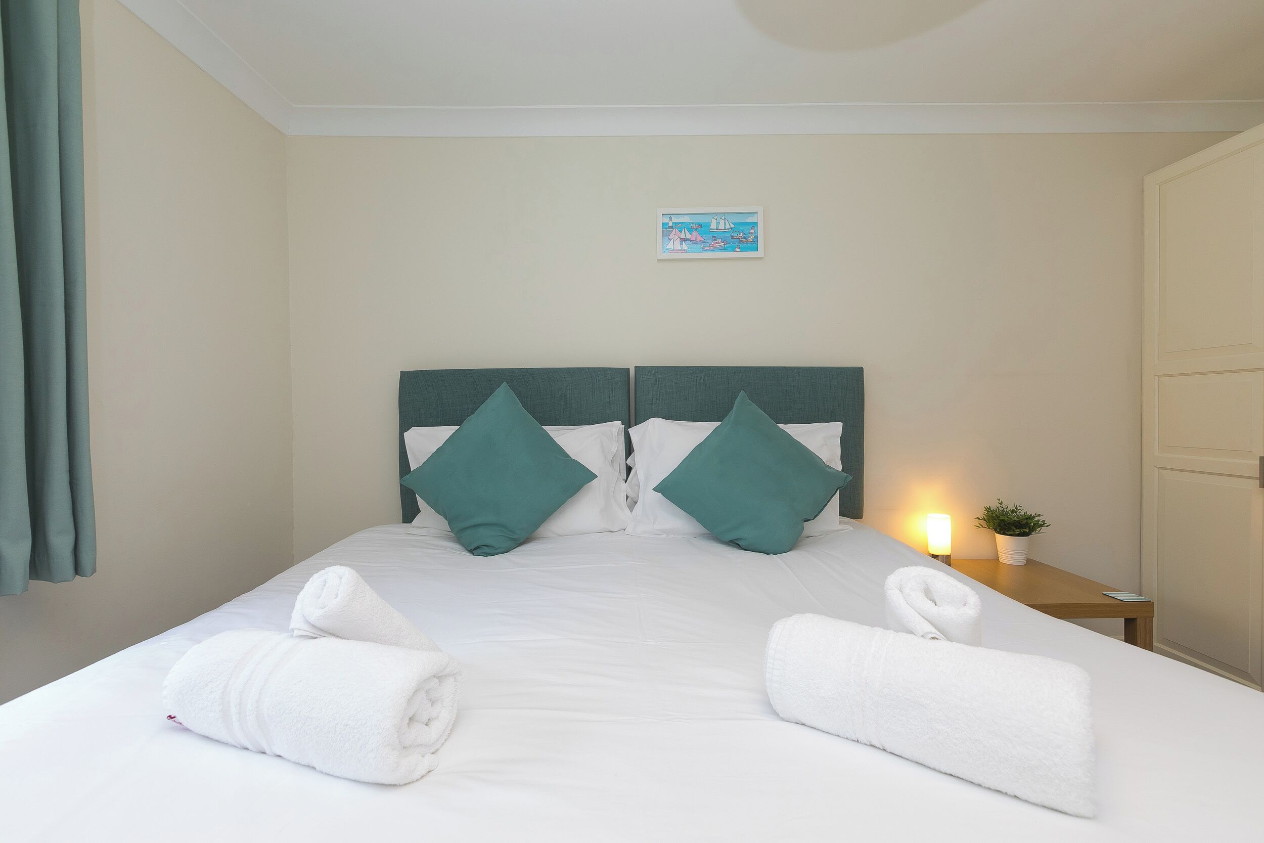 A neatly made bed with two pillows, a bedside table with a lamp and a plant, and a wall-mounted picture.