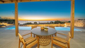 Un espacio de estar al aire libre con techo, piscina, muebles de madera y vistas al atardecer.