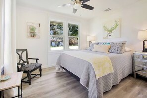 A bedroom with a bed, a chair, a nightstand, a lamp, a ceiling fan, and a window with a view of the outside.