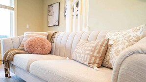 A beige sofa with patterned cushions and a fluffy pillow, positioned in a well-lit room with a window and artwork on the wall. - Designer 3BR Retreat | Steps from Stadiums (Seattle)