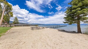 A sandy beach with picnic tables, a wooden pier extending into calm water, and a mountainous backdrop under a partly cloudy sky. - Starry Skies - West Shore Cabin.  Walk to Private Beach. DOG OK (Tahoe City)