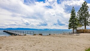 A beach with a wooden pier extending into calm waters, a picnic table, and a tall evergreen tree. - Starry Skies - West Shore Cabin.  Walk to Private Beach. DOG OK (Tahoe City)