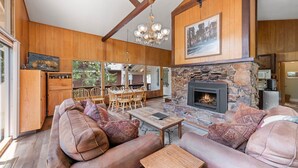 A spacious living room with a stone fireplace, wooden beams, and a large window overlooking a deck. - Starry Skies - West Shore Cabin.  Walk to Private Beach. DOG OK (Tahoe City)