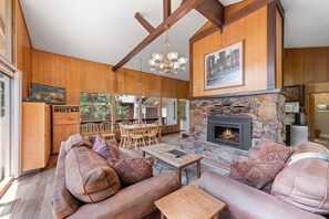A spacious living room with a stone fireplace, wooden beams, and a large window overlooking a deck.