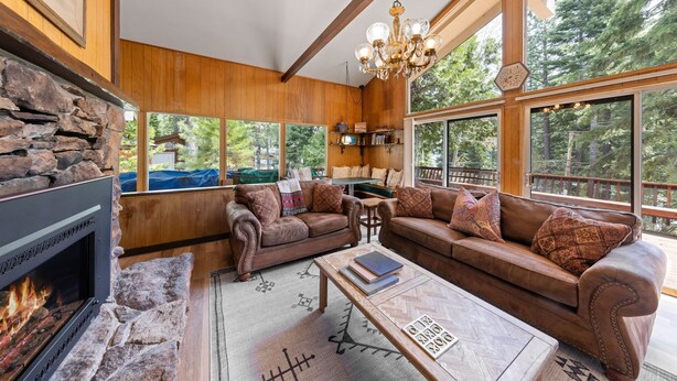 A cozy living room with a stone fireplace, leather sofas, and large windows offering a view of the forest. - Starry Skies - West Shore Cabin.  Walk to Private Beach. DOG OK (Tahoe City)