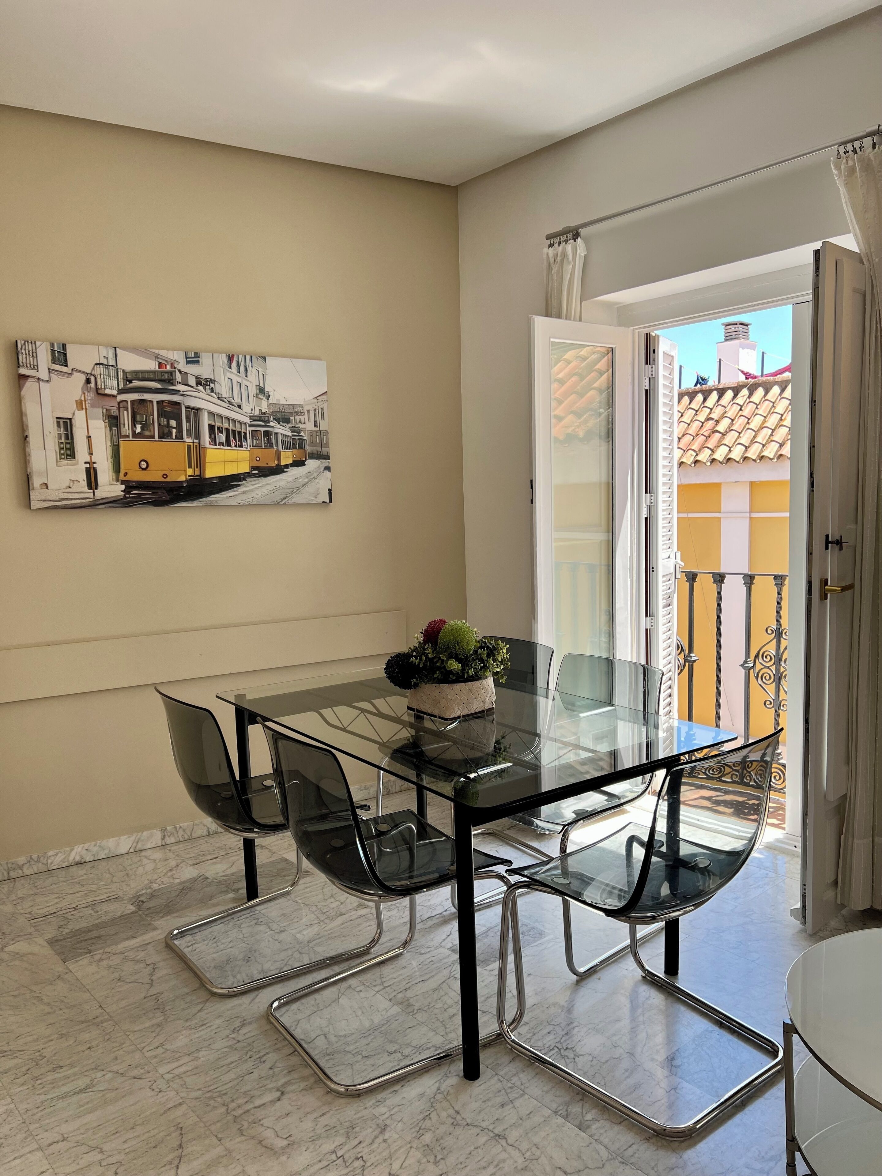 A dining area with a glass table and chairs, a potted plant, and a cityscape painting on the wall.