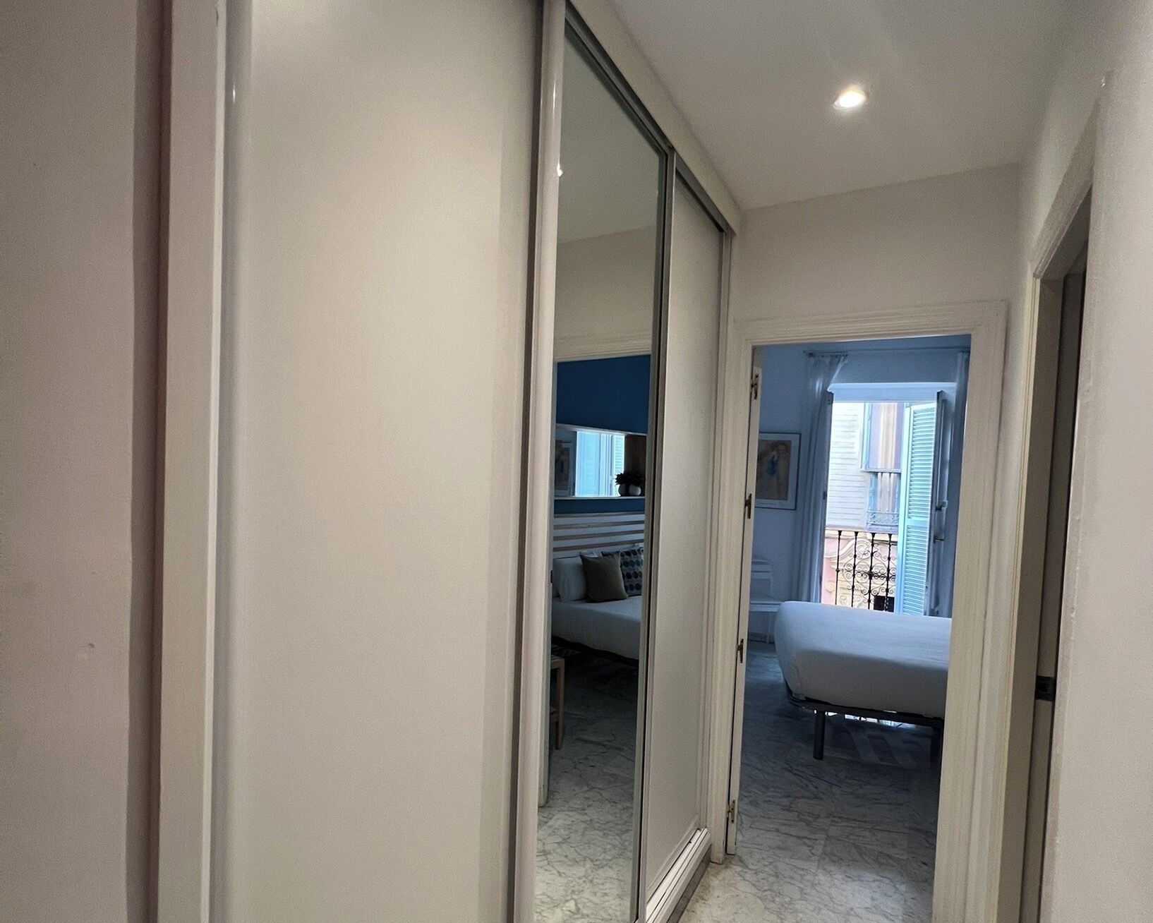 A hallway leading to a bedroom with a large mirror, a bed, and a view of the outside balcony.