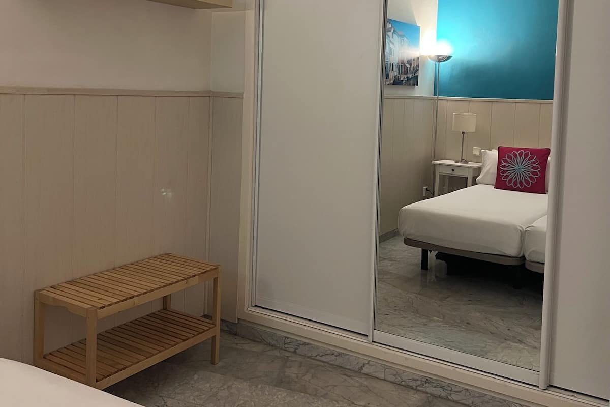 A hotel room with a wooden bench, a bedside table with a lamp, a bed with a red pillow, and a mirror reflecting a cityscape.