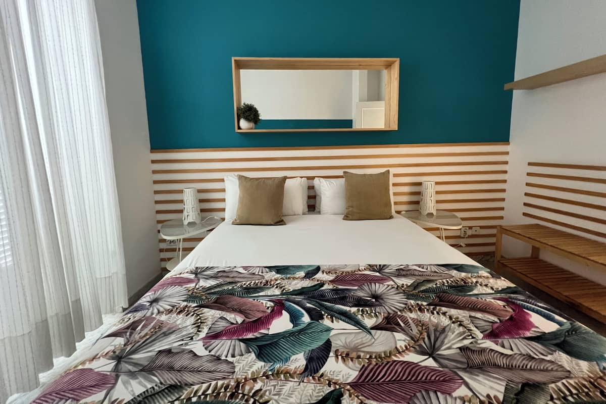 A hotel room with a bed, a bedside table, a mirror, and a window with curtains.