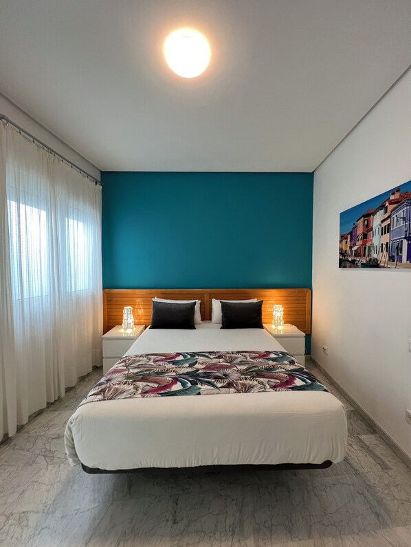 Un dormitorio con cama, dos mesitas de noche, cabecero, ventana con cortinas y un cuadro en la pared.