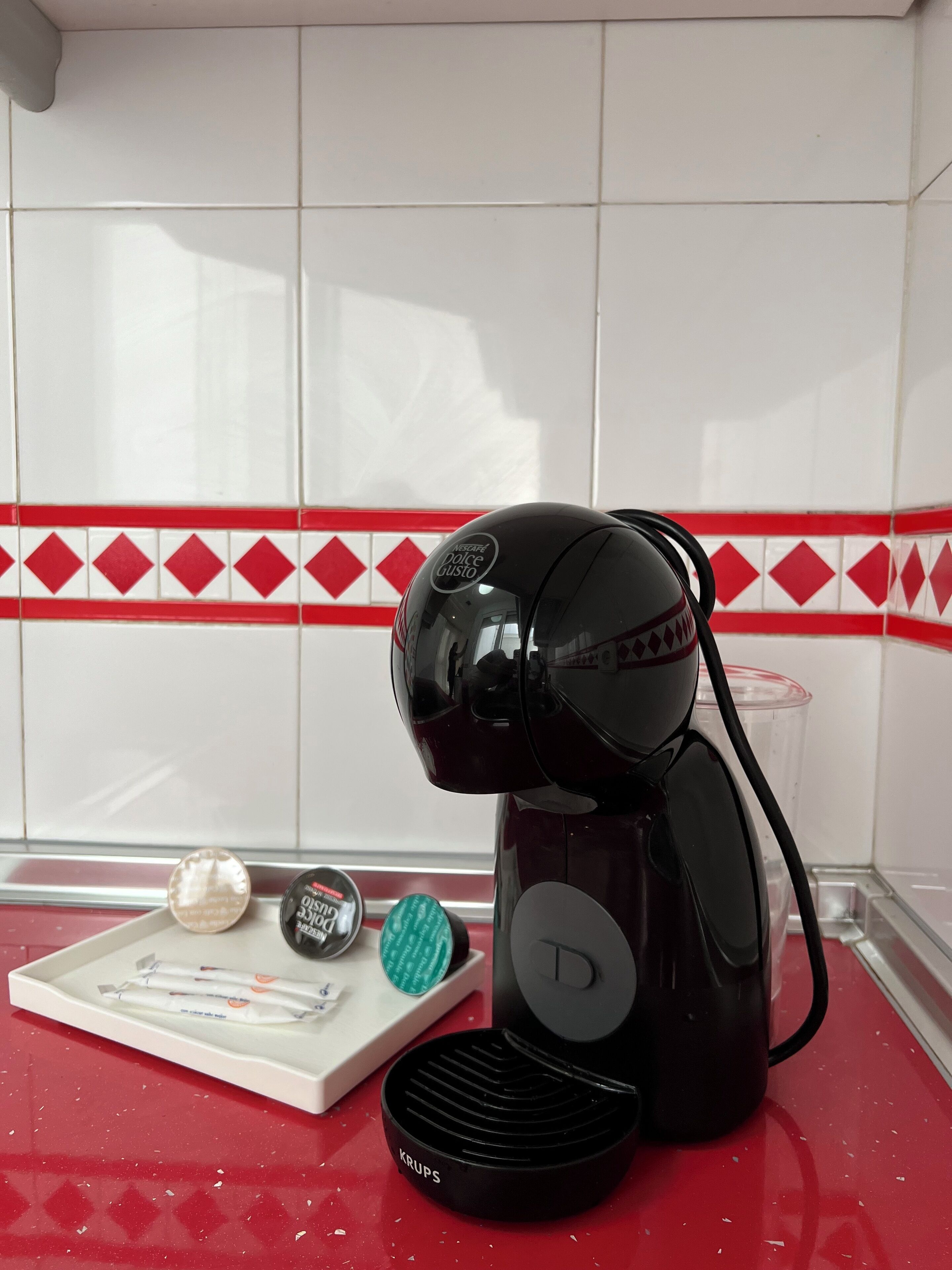 A coffee maker on a red countertop with a tray of coffee pods, a spoon, and a container of sugar.
