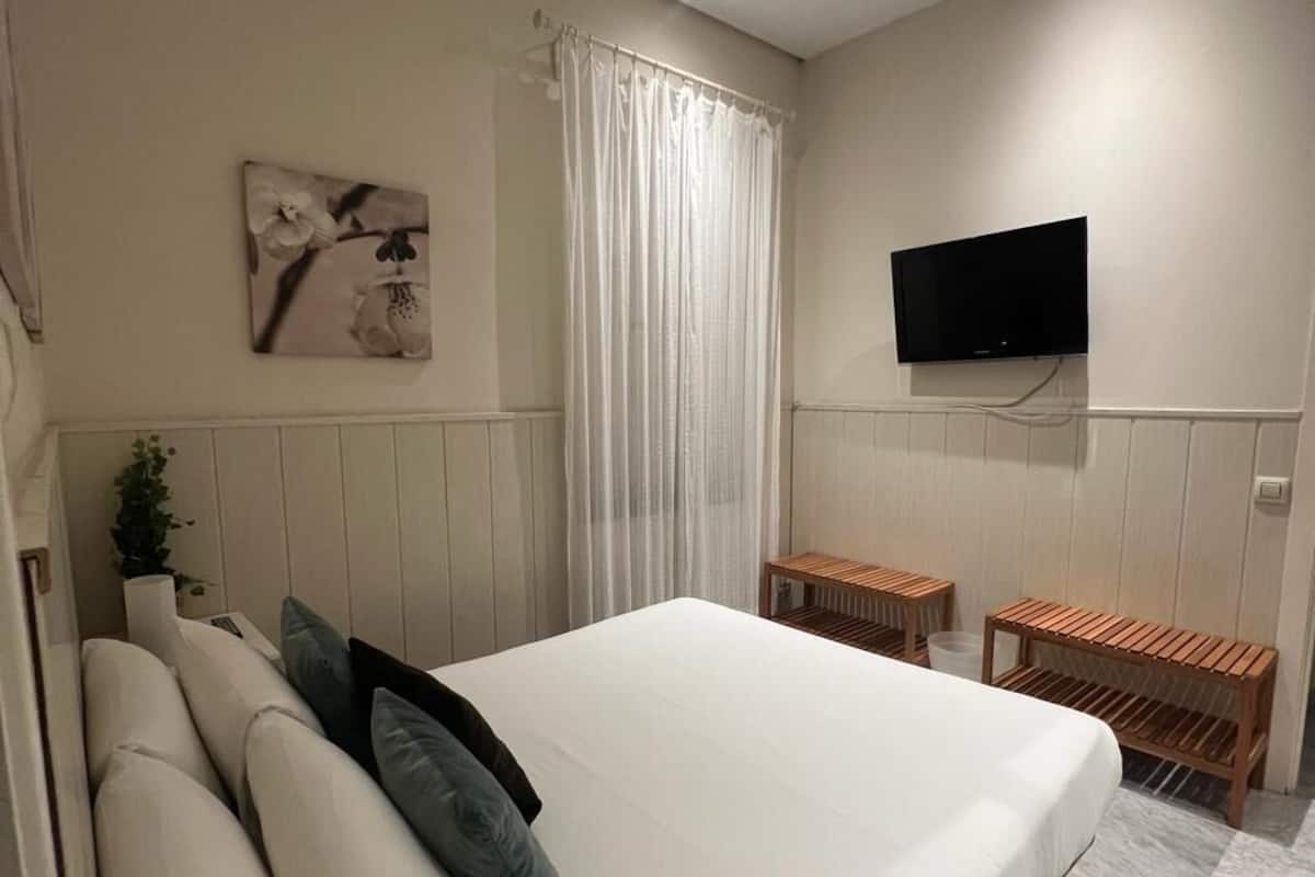 A bedroom with a bed, a TV mounted on the wall, a window with curtains, and a small wooden table.