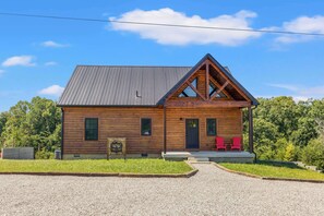 A wooden cabin with a sloped roof, a porch with red chairs, and a gravel driveway.