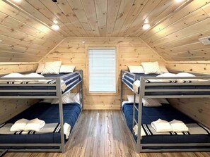 A small attic bedroom with two bunk beds, wooden walls, a window, and a ceiling fan.
