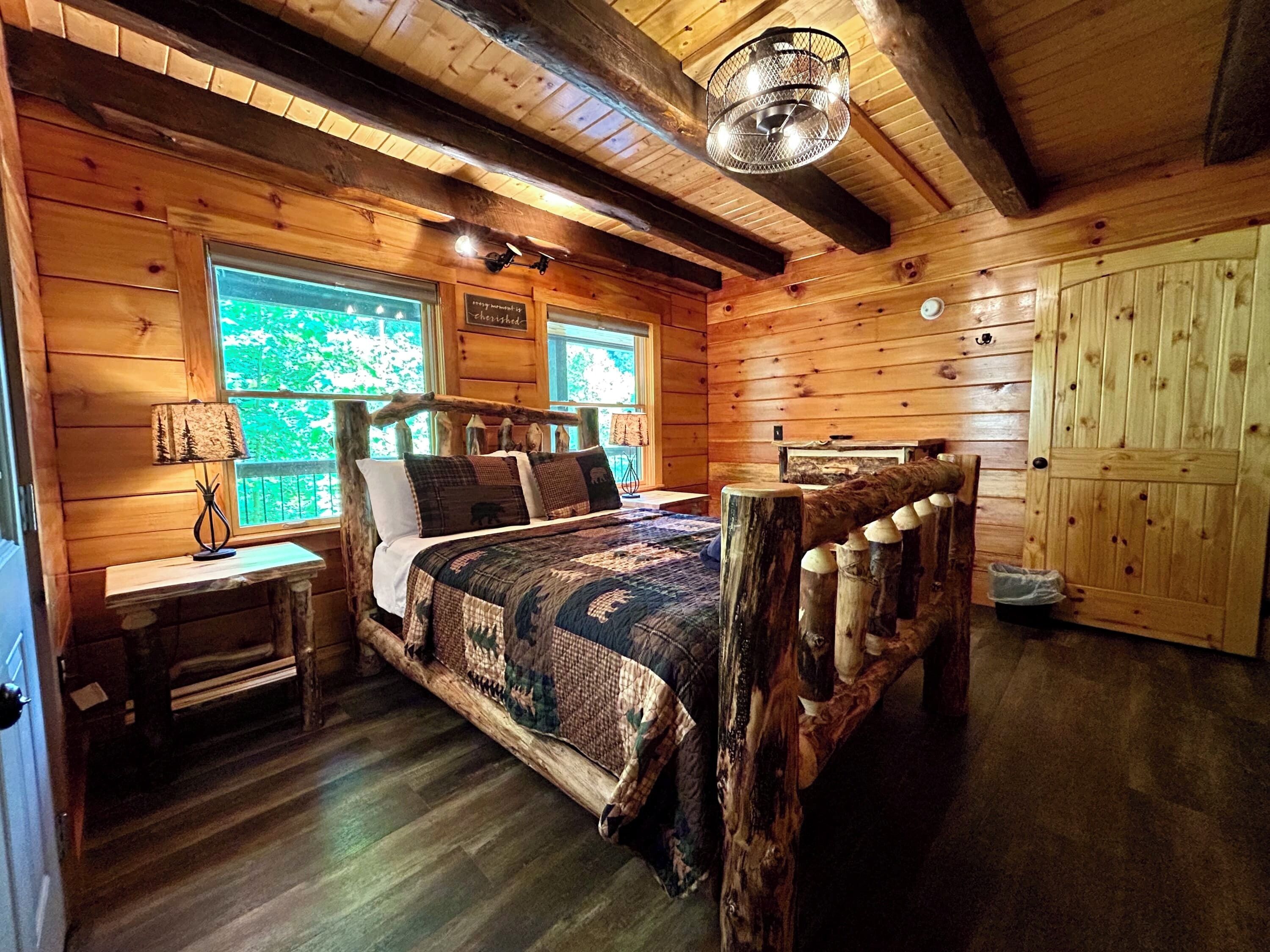 Une chambre à coucher située dans une cabane en rondins, avec un lit, une table de chevet, une lampe et une fenêtre donnant sur la forêt.