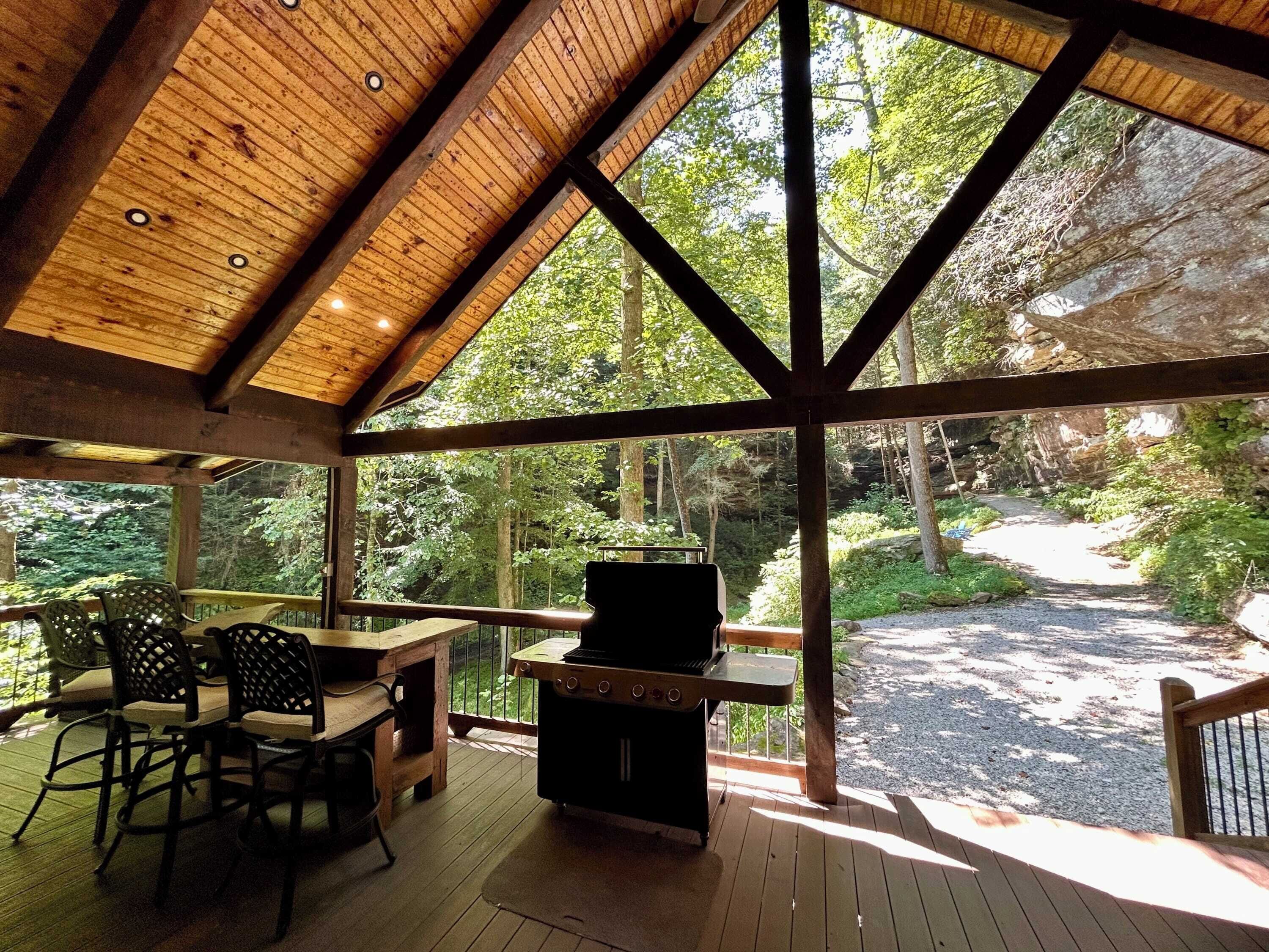 A covered outdoor area with a grill, seating, and a view of a forested area.