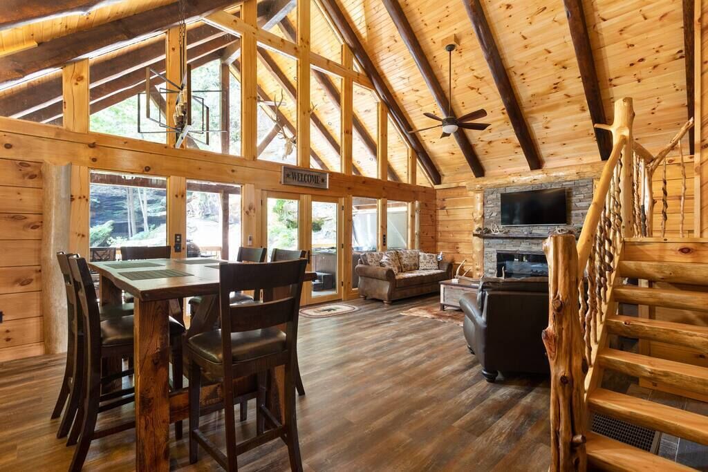 A spacious wooden cabin with a dining area, a living room with a fireplace, and a large window overlooking the forest.