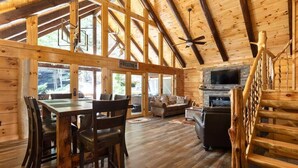 A spacious wooden cabin with a dining area, a living room with a fireplace, and a large window overlooking the forest. - Cabin of Freedom Falls -Private Waterfall, Hot Tub- Secluded & Luxury Log Cabin Retreat (Slade)