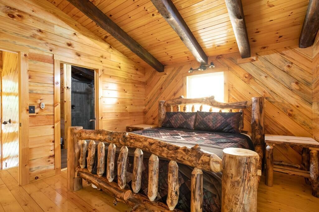 A log cabin bedroom with a wooden bed, a rustic bench, and a door leading to another room.