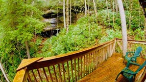 A wooden deck with a railing, surrounded by dense greenery and a small waterfall. - Cozy RRG Retreat | Private Valley & Forest Views | Hidden Falls Cabin (Campton)