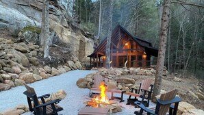 Eine Lodge mit Kamin aus Stein, umgeben von Adirondack-Stühlen, vor Waldkulisse.