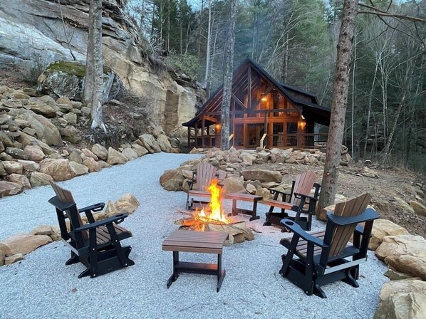 Un chalet doté d’une cheminée en pierre, entouré de fauteuils Adirondack, le tout se fondant dans un décor forestier.