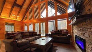 Ein geräumiges Wohnzimmer mit hohen Holzbalken, großen Fenstern, einem Kamin aus Stein und Ledersofas.