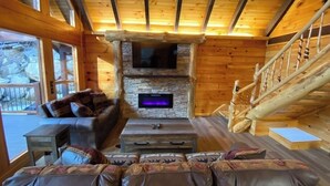 Ein Blockhaus-Interieur mit Kamin aus Stein, einem Wand-Fernseher, einer Holztreppe und einer Couch mit gemusterten Kissen.