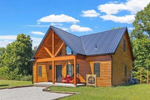 A log cabin with a pitched roof, a porch with red chairs, and a sign with a mountain design. - Hilltop Hideaway-NaturalBridge2m-Modern-HotTub (Rogers)