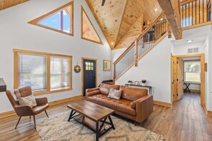 A spacious living room with a high ceiling, wooden beams, a leather sofa, a wooden coffee table, and large windows.