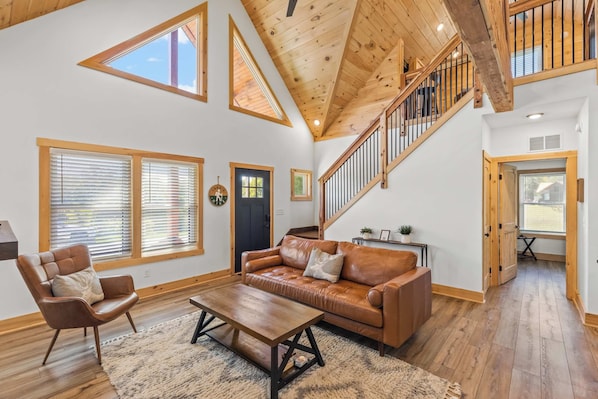 A spacious living room with a high ceiling, wooden beams, a leather sofa, a wooden coffee table, and large windows.