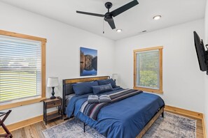 A bedroom with a bed, a ceiling fan, a nightstand, a lamp, and a painting on the wall. - Hilltop Hideaway-NaturalBridge2m-Modern-HotTub (Rogers)