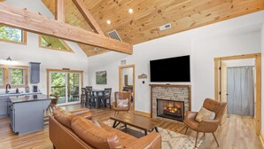 A spacious living area with a fireplace, wooden ceiling beams, a large TV, and a dining area with a view of the outdoors.