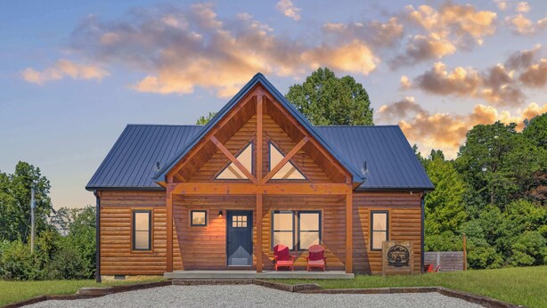 A log cabin with a gabled roof, a front porch with two red Adirondack chairs, and a gravel driveway.