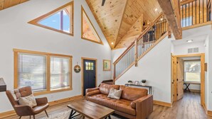 A spacious living room with a high ceiling, wooden beams, a leather sofa, a wooden coffee table, and large windows.