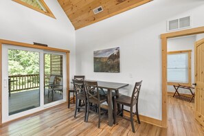 A dining area with a table and chairs, a large window with a view of greenery, and a wooden ceiling. - Hilltop Hideaway-NaturalBridge2m-Modern-HotTub (Rogers)