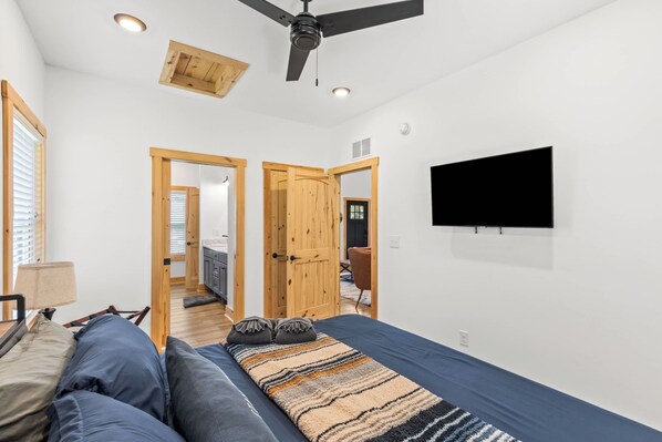 A bedroom with a bed, a TV mounted on the wall, a ceiling fan, and wooden doors leading to other rooms.