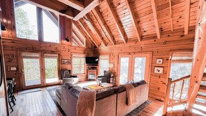 A cozy cabin interior with a wooden floor, a central seating area with a sofa and armchairs, a television, and a fireplace. - Horseshoe Falls-RRG-Hot tub! (Rogers)