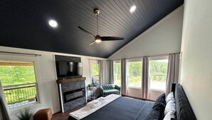 Quarto moderno com cama grande, ventilador de teto, lareira, TV e varanda com vista para área verde.