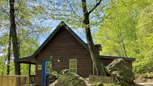 Una baita di legno con una porta blu, circondata da alberi e un grande masso.