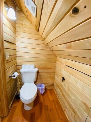 Ein kleines Badezimmer mit Holzwandverkleidung, einer Toilette, einem Mülleimer und einem Lüftungsgitter an der Decke.