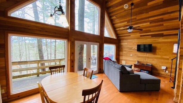 A spacious cabin with a wooden interior, large windows, a dining area, a sofa, and a ceiling fan. - Mountain Chalet |Hot Tub | Centrally Located | RRG (Campton)