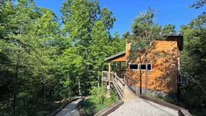 A wooden cabin with a wooden deck and a gravel path leading to it, surrounded by dense forest.
