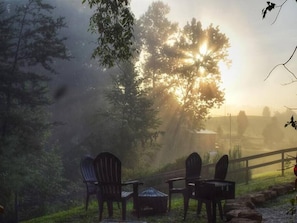 Un espacio de estar al aire libre rústico con sillas Adirondack y una mesa pequeña, rodeado de árboles y una valla de madera.